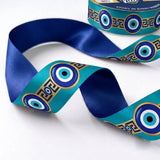 Close-up of Nazar Key ribbon: evil eyes along a gold Greek key band printed on teal satin, royal blue back